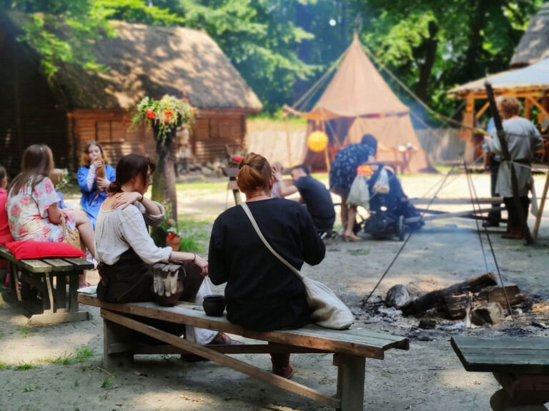 Ludzie siedzący przy ognisku pośrodku zrekonstruowanej wczesnośredniowiecznej osady.
