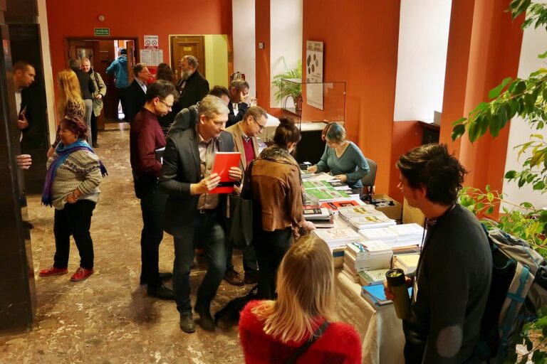 Zdjęcie przedstawia ludzi w holu muzeum. Niektórzy rozmawiają, inni podchodzą do stołu z książkami poświęconymi archeologii.