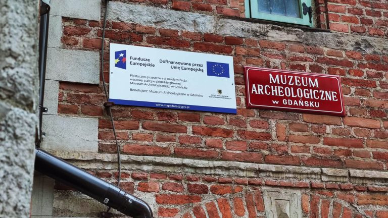 Fragment ceglanej ściany muzeum z dwiema tablicami. Na jednej jest nazwa Muzeum Archeologiczne w Gdańsku, na drugiej informacja o dofinansowaniu przez Fundusze Europejskie.