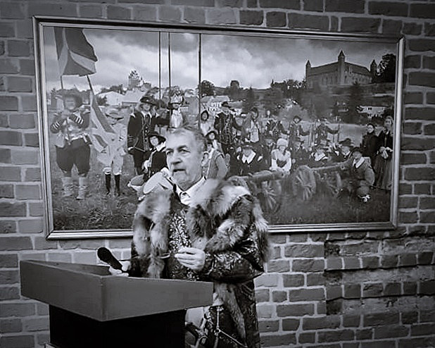 Zdjęcie mężczyzny ubranego w szlachecki kontusz, stojącego przy pulpicie, na tle obrazu.