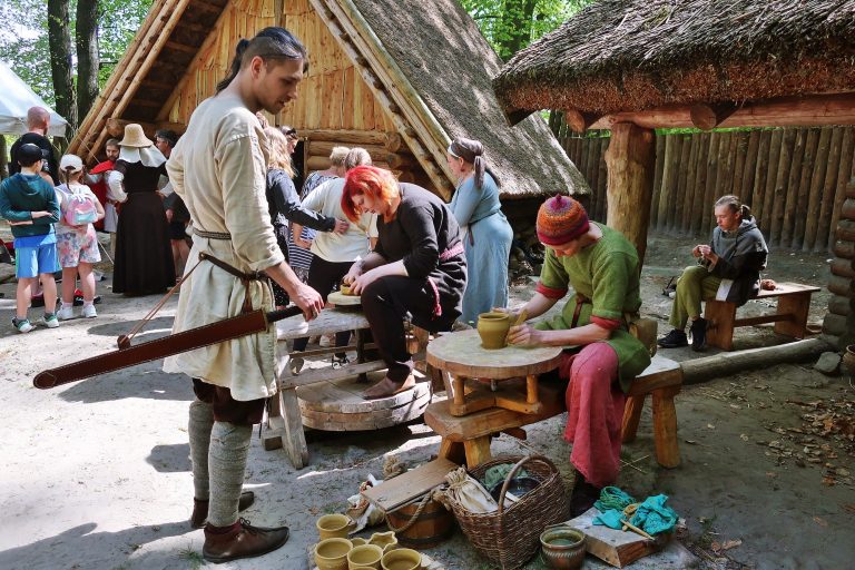Zdjęcie przedstawiające warsztaty garncarskie na sopockim grodzie. W tle drewniana chata i stojący przed nią ludzie. Na pierwszym planie kobieta w kolorowym stroju z epoki średniowiecza, pracująca przy kole garncarskim, obok niej stoi mężczyzna w średniowiecznym stroju z mieczem przy boku.