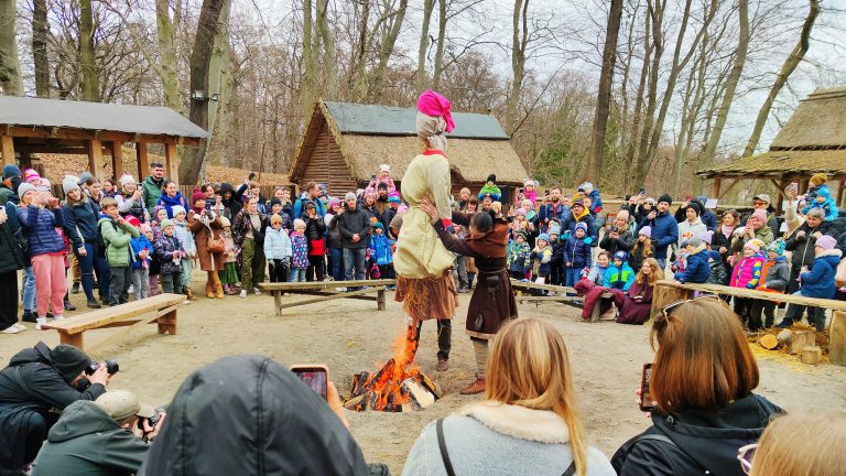 Grodzisko w Sopocie, pośrodku płonie ognisko, przy ognisku stoi mężczyzna, który wkłada do ogniska dużą, kolorową kukłę-. Dookoła ogniska są ustawione ławy, za nimi stoją ludzie, przyglądają się przedstawieniu.